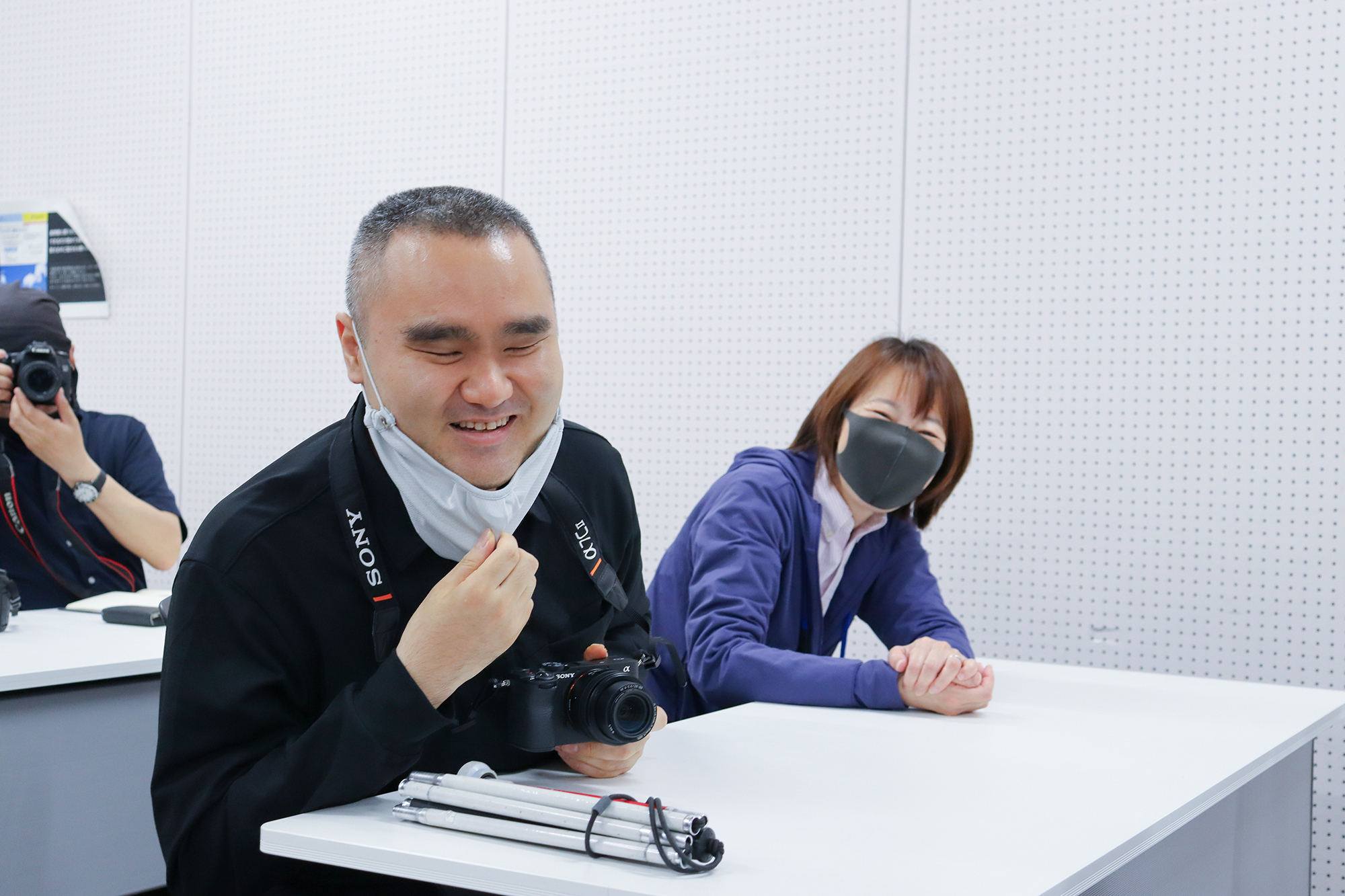 マスクを少し下げ、さらに笑顔を見せる濱崎さんとガイドさん。机の上にはカメラと白杖が置かれている。
