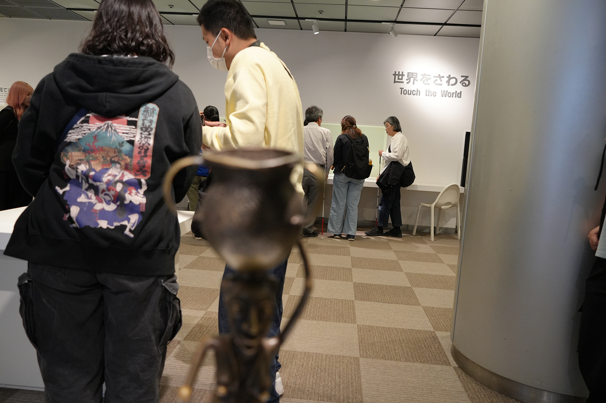 真鍮製のミム像を手前にぼかし、奥で展示を見る安田さんたちにピントを合わせた展示室内の写真。