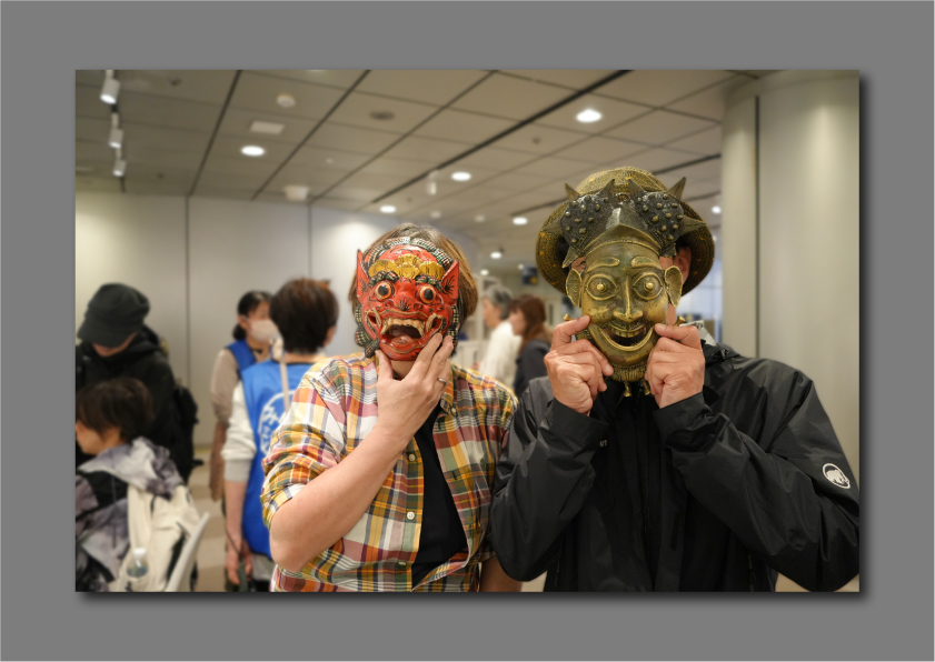 二人の人物が真鍮製と木製の仮面を被って楽しそうに立っている作品