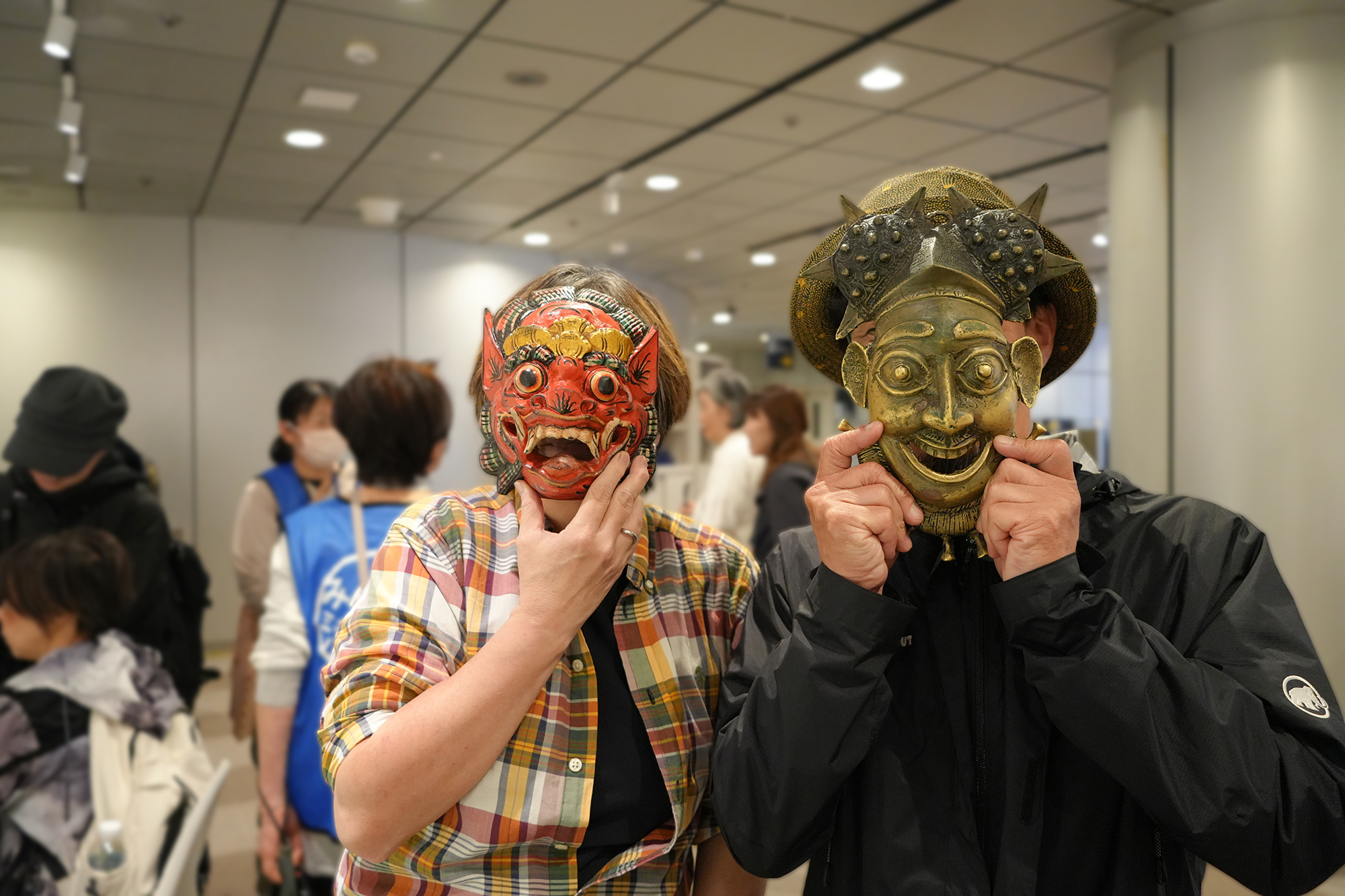 二人が仮面をつけて並ぶ様子を撮影した写真。背景は少しぼけ、人物が際立っている。